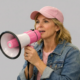 Woman wearing a pink cap speaking into a pink megaphone against a neutral background.