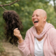 Bald woman laughing outdoors while holding a wig, representing humor and resilience during cancer treatment.